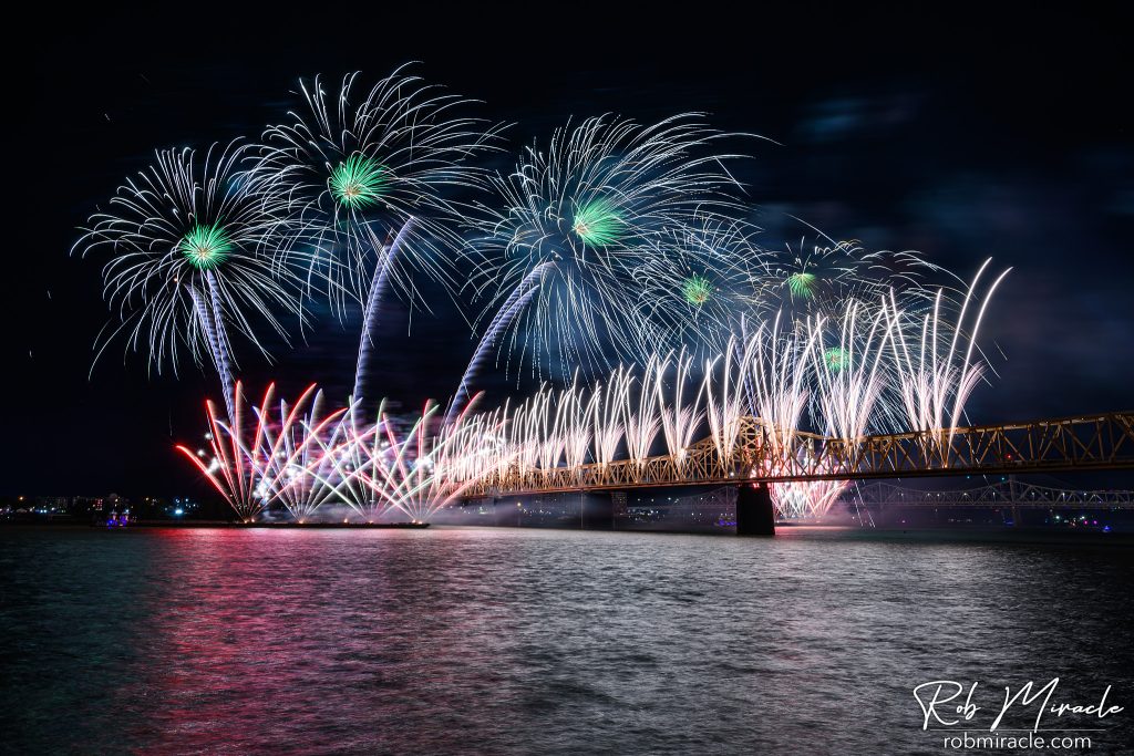 Fireworks launching from a barge in a river and from a bridge crossing the river.