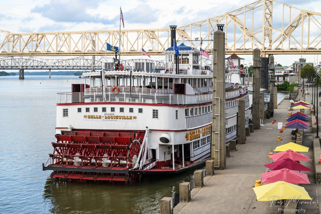 A view of the historic Belle of Louisville from behind while docked at the wharf. A bridge and river can be seen behind the boat and colorful umbrellas line the right side of the wharf.
