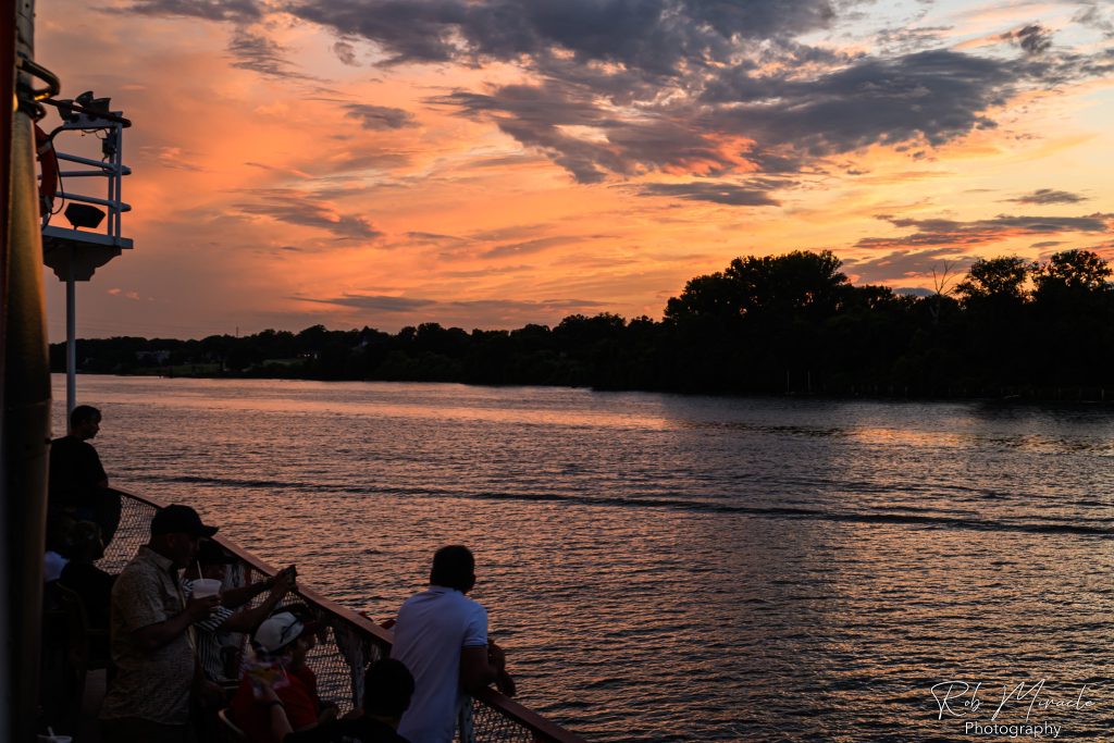 A silhouetted passenger looks right off of the bow of a steamboat as the sunset over the Ohio River.