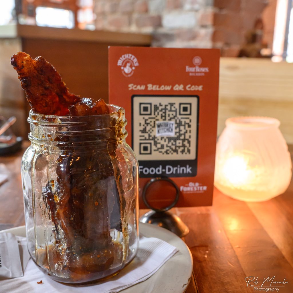 Several strips of maple candied bacon sit in a glass mason jar to the left, while the QR code to scan Merle's menu is in the center with a candle on the right.