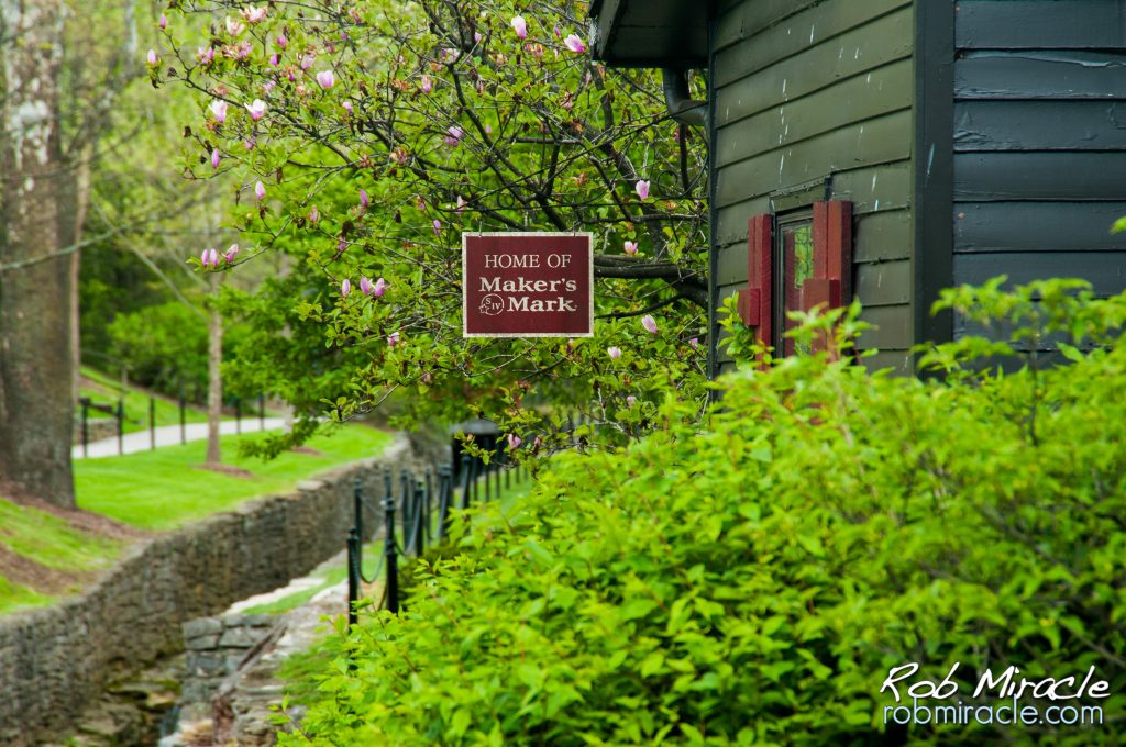 Green scenery with a grey building with red trim with a sign that reads "Makers Mark" hanging from the side.